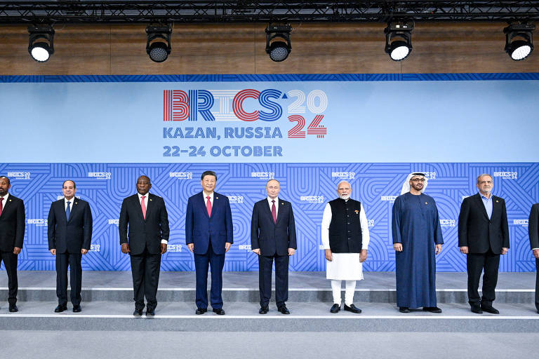 A imagem mostra um grupo de líderes de diferentes países posando em um evento do BRICS 2024. Eles estão alinhados em uma plataforma， com um fundo que exibe o logotipo do evento e as datas de 22 a 24 de outubro. Os líderes estão vestidos de maneira formal， com trajes típicos de seus países. O ambiente é bem iluminado e organizado.