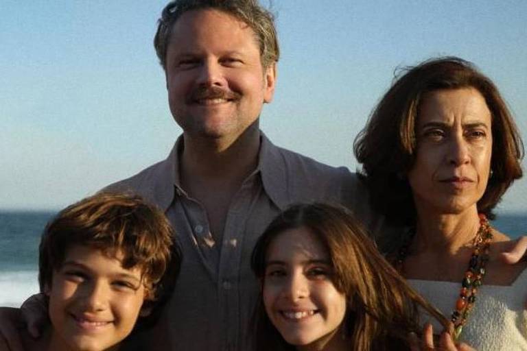 Uma família posando em frente ao mar. Um homem com bigode e cabelo grisalho está no centro， sorrindo. À sua esquerda， um menino com cabelo castanho claro e ondulado， e à sua direita， uma menina com cabelo castanho escuro e liso. Uma mulher com cabelo castanho e liso， usando uma blusa clara， está ao lado do homem， com uma expressão séria. O fundo mostra o mar e o céu.