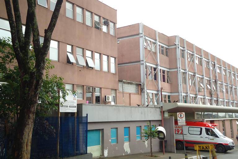 Imagem de um edifício de hospital com uma estrutura de dois andares. A fachada é de cor marrom e possui janelas com algumas abertas. Na parte inferior， há uma entrada com um toldo e uma ambulância estacionada. À esquerda， há uma árvore e uma grade azul. O céu está nublado.