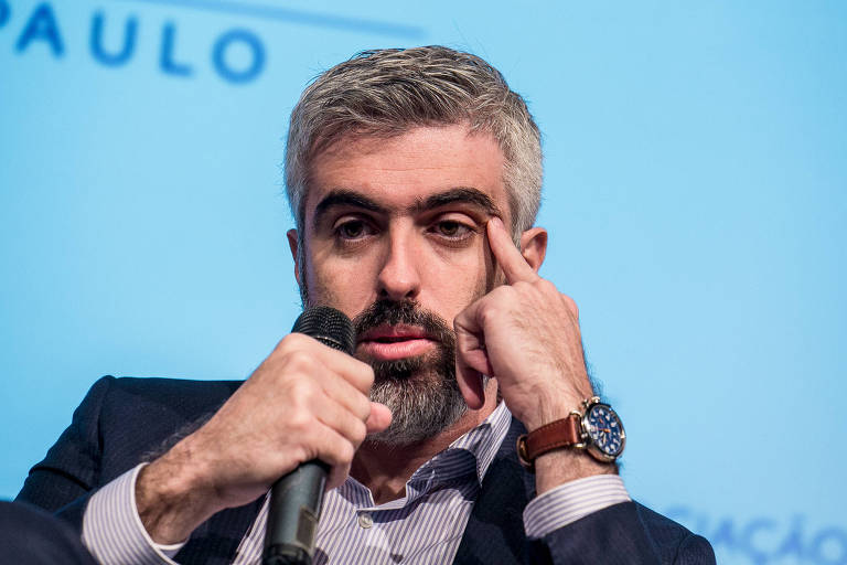 Um homem com cabelo curto e grisalho está sentado em um palco， segurando um microfone com a mão esquerda. Ele está olhando para a frente e tocando a parte de seu rosto com a mão direita， em uma expressão pensativa. O fundo é azul claro