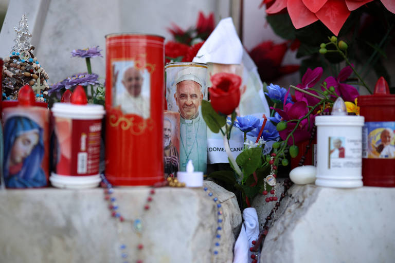 A imagem mostra um altar com velas acesas, flores e imagens religiosas. Há velas vermelhas e brancas, algumas com fotos do Papa Francisco. Também estão presentes flores coloridas e um lenço branco. O ambiente é decorado com contas e outros elementos religiosos.