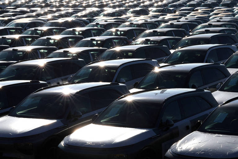 A imagem mostra um grande estacionamento repleto de carros， predominantemente de cor prata. Os veículos estão organizados em filas， refletindo a luz do sol， o que cria um efeito brilhante na superfície dos carros. A cena é ampla e parece não ter fim， com muitos carros visíveis ao fundo.