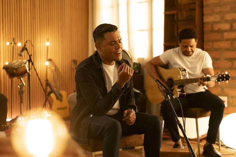 A imagem mostra dois homens em um ambiente de estúdio musical. Um deles está sentado em uma cadeira, cantando com uma expressão emocional, enquanto o outro está ao fundo, tocando violão. O cenário é acolhedor, com paredes de madeira e iluminação suave. Instrumentos musicais estão visíveis ao fundo.