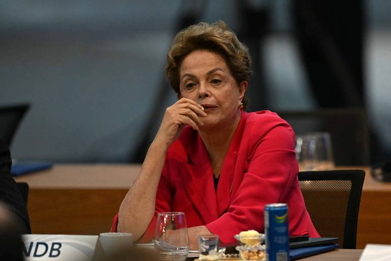 Imagem mostra Dilma Rousseff， uma mulher sentada em uma mesa de reunião， com uma expressão pensativa. Ela usa um blazer rosa e está com a mão no queixo. Na mesa， há copos e um recipiente com comida. Ao fundo， há uma mesa com documentos e outros participantes da reunião.