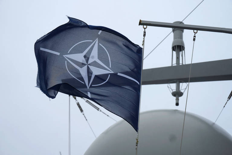 A imagem mostra a bandeira da Otan (Organização do Tratado do Atlântico Norte) tremulando ao vento. A bandeira é de cor azul escura com um emblema central que representa uma estrela de quatro pontas， cercada por um círculo. Ao fundo， há uma estrutura de um navio， com uma esfera branca visível.