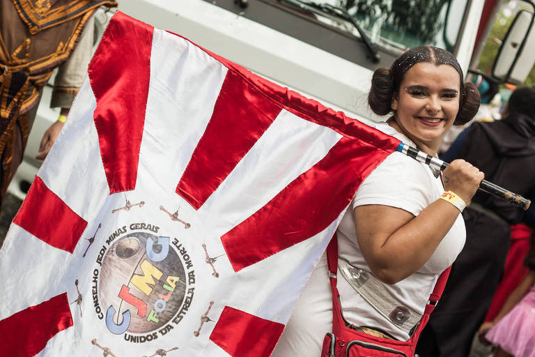 Uma mulher sorridente segura uma bandeira com um design em vermelho e branco. A bandeira possui um emblema central que diz 039;GIMC039; e outras inscrições. A mulher está vestida com um traje claro e tem cabelo preso em coques. Ao fundo， há outras pessoas e um veículo.