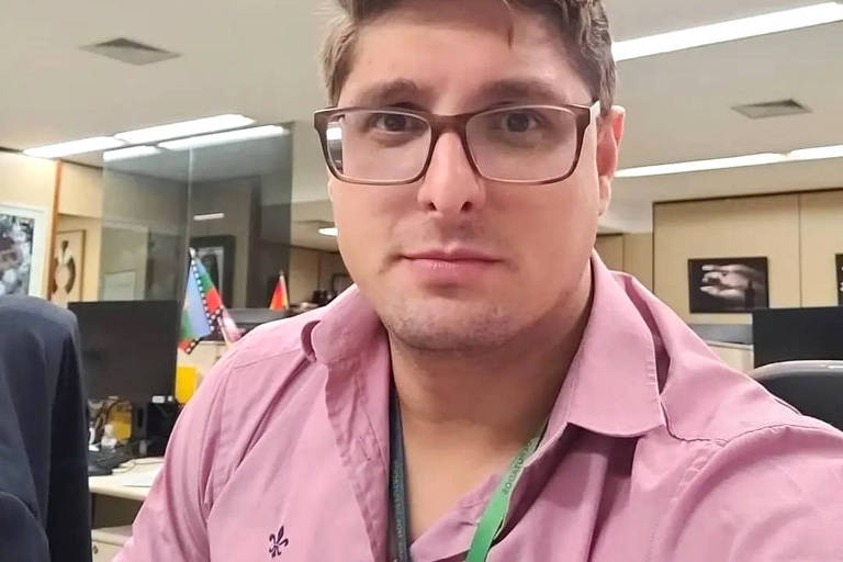 Um homem com cabelo castanho claro e óculos está posando para a câmera em um ambiente de escritório. Ele usa uma camisa rosa e um crachá verde. Ao fundo, há mesas de trabalho e algumas bandeiras decorativas.