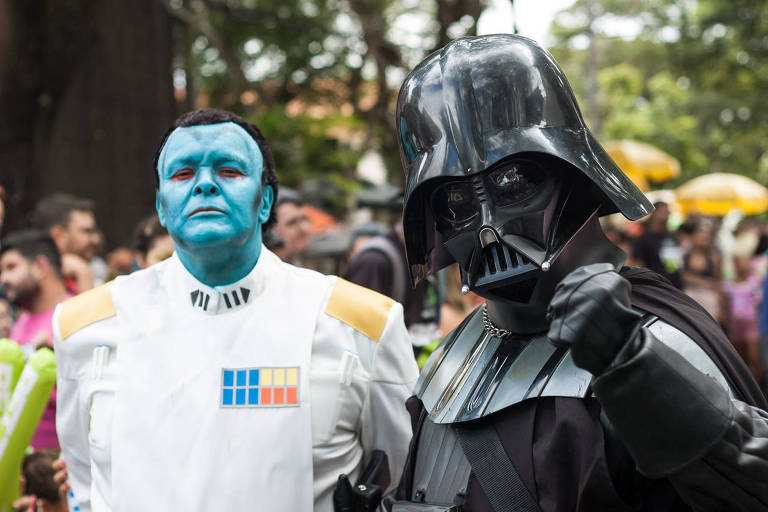 A imagem mostra duas pessoas vestidas como personagens do universo Star Wars. À esquerda， um homem com a pele azul， usando um uniforme branco com detalhes em amarelo e azul. À direita， um homem vestido como Darth Vader， com um traje preto e capacete característico， fazendo um gesto de punho cerrado. Ao fundo， há uma multidão e algumas árvores.