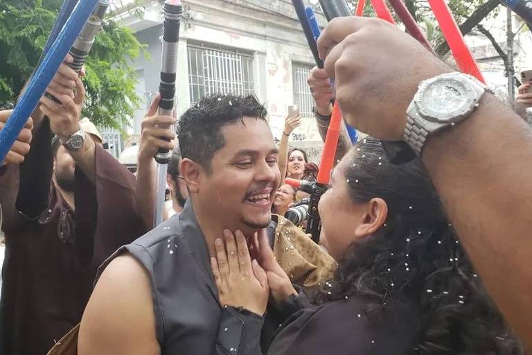 A imagem mostra um grupo de pessoas em uma celebração ao ar livre. No centro， um homem sorridente está interagindo com uma mulher， que o segura pelo rosto. Ao redor deles， várias pessoas seguram bastões coloridos， e há uma atmosfera festiva com confetes ou pétalas caindo. O cenário parece ser um evento alegre， possivelmente um casamento ou uma festa.