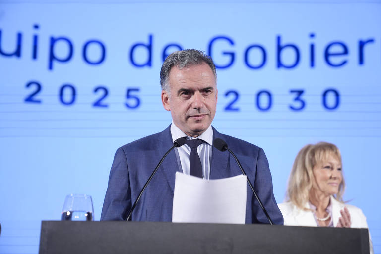 Um homem em um terno escuro está em pé em um palco， segurando um papel e falando ao microfone. Ao fundo， há um grande painel com o texto 039;Equipo de Gobierno 2025-2030039;. À direita， uma mulher com cabelo loiro e vestido claro observa. Uma taça de vidro está sobre a mesa à frente do homem.