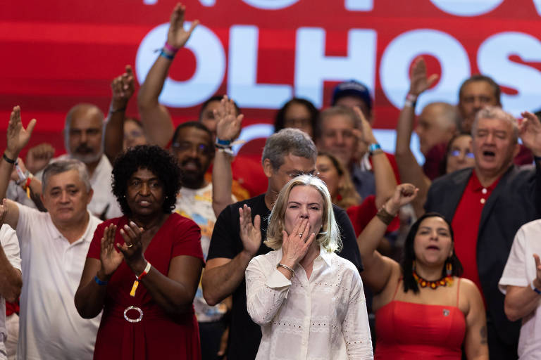 A imagem mostra uma multidão em um evento político. No centro， uma mulher de cabelo liso e claro， vestindo uma blusa branca， está fazendo um gesto de beijo. Ao fundo， várias pessoas aplaudem e levantam as mãos， algumas vestindo roupas vermelhas e brancas. O fundo é vermelho com texto em letras grandes que diz 039;NOTÓRIO OLHOS039;.