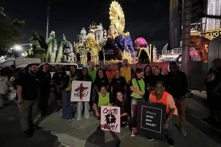 Um grupo de pessoas posando para a foto em um evento noturno, com um carro alegórico iluminado ao fundo. Algumas pessoas estão segurando cartazes com as inscrições 'Cristo Vive!' e 'Posso orar por vocês?'. O ambiente é festivo, com luzes coloridas e decoração típica de celebrações