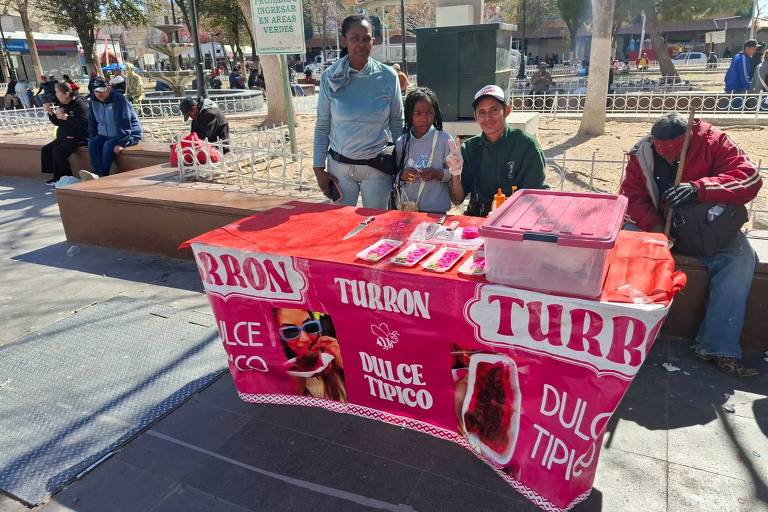 A imagem mostra uma banca de doces em uma praça. A mesa é coberta com uma toalha vermelha e branca， com as palavras 039;TURRÓN039; e 039;DULCE TÍPICO039; em destaque. Atrás da mesa， há três pessoas: um homem e uma mulher adultos， e uma criança. O ambiente é ao ar livre， com outras pessoas visíveis ao fundo， sentadas em bancos.