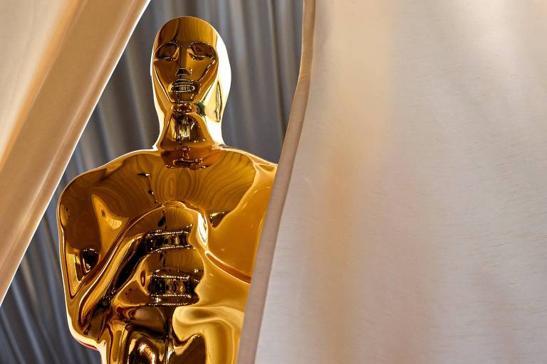 A imagem mostra uma estátua dourada do Oscar, parcialmente visível entre cortinas claras. O fundo é de um tom neutro, destacando a figura brilhante da estatueta.