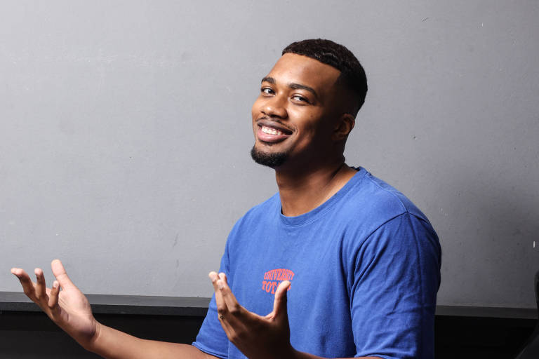 Um homem está sentado em uma cadeira preta, vestindo uma camiseta azul e shorts. Ele tem um sorriso no rosto e está gesticulando com as mãos, como se estivesse explicando algo. O fundo é cinza, e a iluminação é suave.