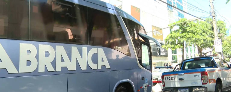 A imagem mostra um ônibus da Viação Itapemirim， de cor azul， estacionado em uma rua. O ônibus possui a palavra 039;BRANCA039; em letras grandes e brancas na lateral. Ao lado， há uma caminhonete de cor azul e branca. Ao fundo， é possível ver um edifício com janelas e uma vegetação ao redor.