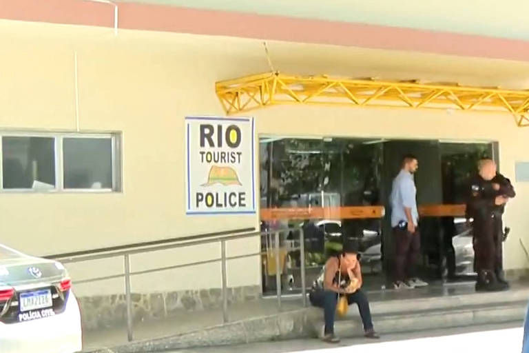 A imagem mostra a entrada de uma delegacia de polícia turística no Rio de Janeiro. Há um letreiro grande com as palavras 039;RIO TOURIST POLICE039;. Na frente， um homem sentado e dois policiais em pé， um deles em uniforme. Um carro da polícia está estacionado próximo à entrada.