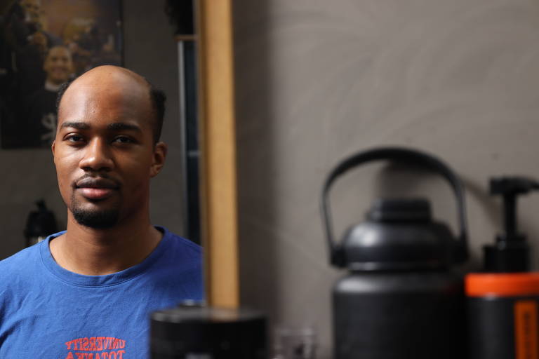 Um homem com cabelo raspado e uma barba curta está posando em frente a um espelho. Ele usa uma camiseta azul e está em um ambiente que parece ser um salão de beleza, com vários produtos de cabelo visíveis ao fundo. O fundo é levemente desfocado, destacando o homem.