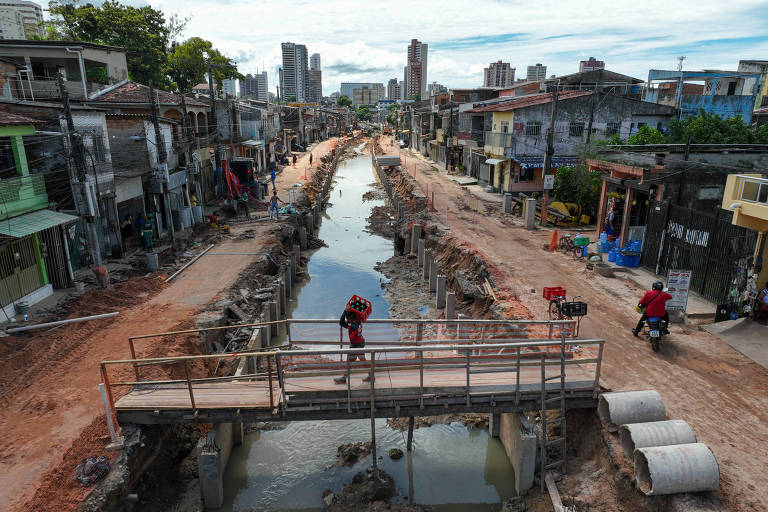 A imagem mostra uma rua em obras， com um canal aberto no meio. Há uma ponte de madeira sobre o canal， e algumas pessoas estão caminhando pela área. Os edifícios ao fundo são de vários andares， e a cena é iluminada por luz natural. O chão está coberto de terra e detritos， e há tubos de concreto ao lado do canal.