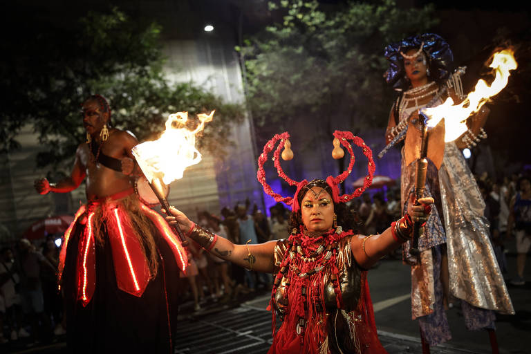 A imagem mostra três performers em um evento cultural noturno. No centro， uma mulher com trajes vermelhos e adornos， segurando tochas acesas. À esquerda， um homem com uma saia vermelha e detalhes luminosos， e à direita， uma mulher com cabelo azul e um vestido brilhante. O fundo é uma multidão e árvores iluminadas.