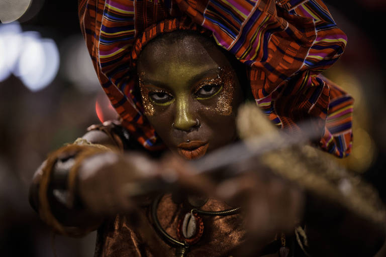 A imagem mostra uma pessoa com maquiagem dourada e brilhante， usando um vestido de cor bronze e um grande turbante colorido. A pessoa está em uma pose que sugere movimento， com um olhar intenso e focado. O fundo é desfocado， com luzes que indicam um ambiente festivo.