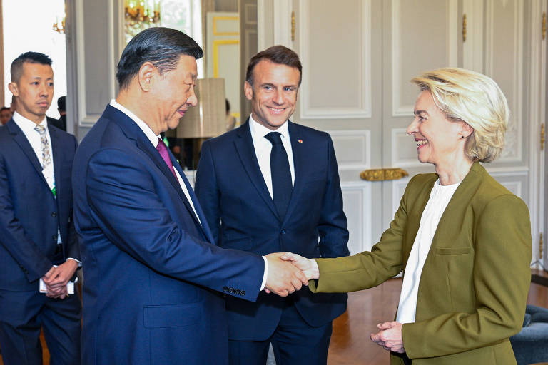 A imagem mostra um encontro entre três pessoas em um ambiente interno elegante. À esquerda， Xi Jinping， de terno escuro， está apertando a mão de von der Leyen， uma mulher de cabelo loiro e curto， que usa um blazer verde. Ao fundo， Macron， um homem de terno escuro， observa com um sorriso. O ambiente é bem iluminado， com paredes claras e detalhes decorativos.