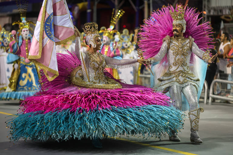 A imagem mostra um casal dançando em um desfile de carnaval. A mulher usa um vestido volumoso com uma saia em tons de rosa e azul, adornado com detalhes dourados. O homem ao seu lado veste um traje brilhante, também com detalhes dourados, e segura uma bandeira. Ao fundo, há outras pessoas fantasiadas, com rostos pintados e trajes coloridos, que fazem parte da mesma apresentação.