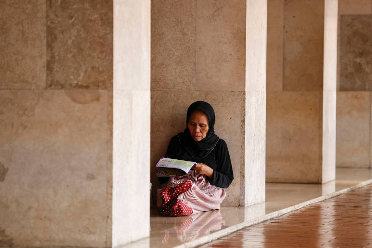 A imagem mostra uma mulher sentada no chão, encostada em uma coluna de um edifício. Ela está vestindo uma roupa escura e um lenço na cabeça, e segura um livro aberto. O ambiente é bem iluminado, com paredes de pedra e um piso brilhante. A mulher parece concentrada na leitura.