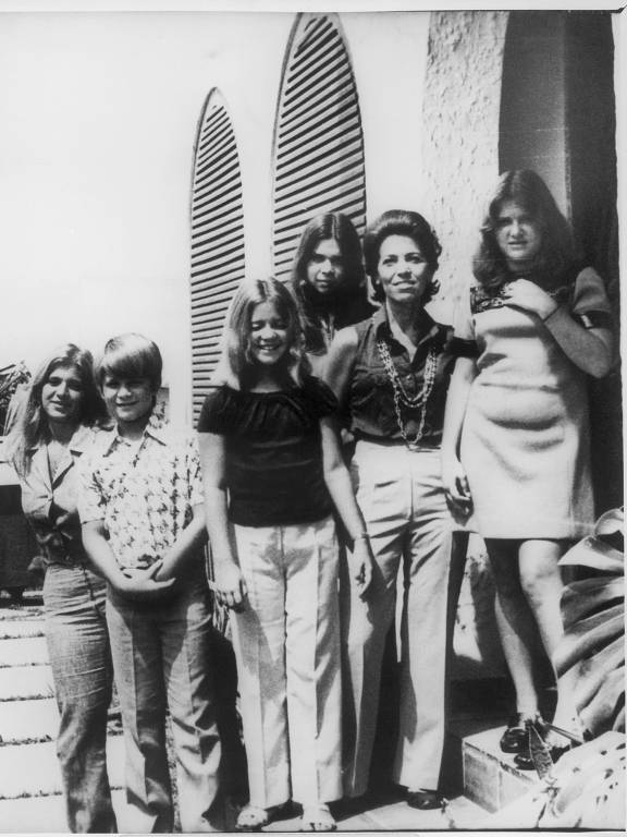 A imagem em preto e branco mostra um grupo de seis pessoas posando em frente a uma parede com janelas arqueadas. Há três mulheres à esquerda, uma criança do meio, e duas mulheres à direita. As mulheres estão vestidas de maneira casual, com uma delas usando uma blusa escura e outra uma saia. A criança está usando uma camisa estampada. A imagem contém uma legenda na parte inferior que menciona a reprodução da família do deputado Rubens Paiva, com a data de 08.12.95 e o nome do fotógrafo Eduardo Simões.