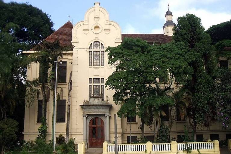 A imagem mostra um edifício histórico de arquitetura clássica， com paredes em tom claro e detalhes ornamentais. O prédio possui um telhado inclinado com uma torre no topo e janelas grandes. À frente， há um jardim com árvores e um muro baixo de cor clara. O céu está parcialmente nublado.