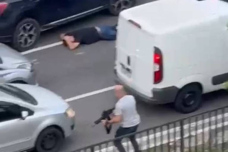 A imagem mostra uma cena tensa em uma via urbana， onde um homem está deitado no chão entre os carros. Um outro homem， segurando uma arma， está em pé próximo a um veículo branco. Vários carros estão parados ao redor， e algumas pessoas parecem estar se agachando ou se escondendo.