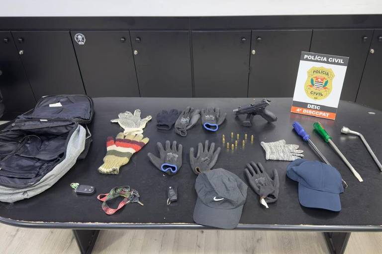 A imagem mostra uma mesa com diversos itens apreendidos pela Polícia Civil. Há uma mochila preta， várias luvas de diferentes tipos， uma arma de fogo， cartuchos， uma chave， um boné e uma barra de metal. Ao fundo， há um cartaz da Polícia Civil com o emblema e a inscrição 039;DEIC039;.