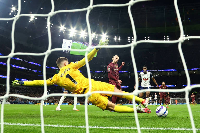 A imagem mostra um momento de um jogo de futebol， onde um goleiro， vestido com um uniforme amarelo， está se esticando para defender um chute. O jogador que está chutando está vestido com uma camisa marrom e parece estar em movimento. Ao fundo， outros jogadores e torcedores são visíveis， com um placar eletrônico ao fundo mostrando o tempo de jogo.