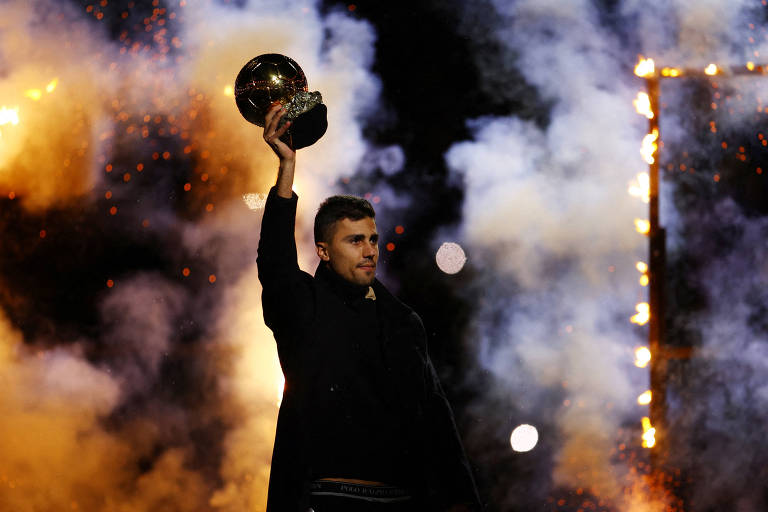 Um jogador de futebol está em um palco， levantando um troféu com uma mão. Ele está vestido com um casaco escuro e tem uma expressão de alegria. Ao fundo， há fumaça e fogos de artifício， criando um ambiente festivo.