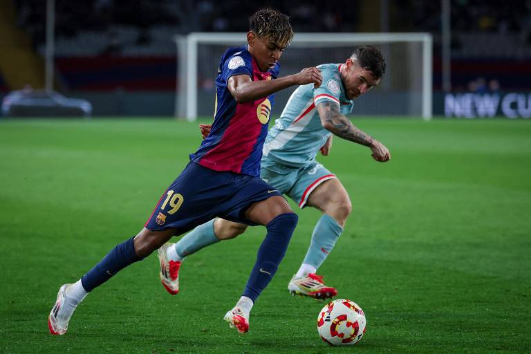 Dois jogadores de futebol estão em campo durante uma partida. Um jogador， vestindo a camisa número 19 do Barcelona， está correndo em direção à bola， enquanto o outro jogador， com uma camisa em tons de azul e branco， tenta alcançá-lo. O campo está bem iluminado e há espectadores ao fundo.