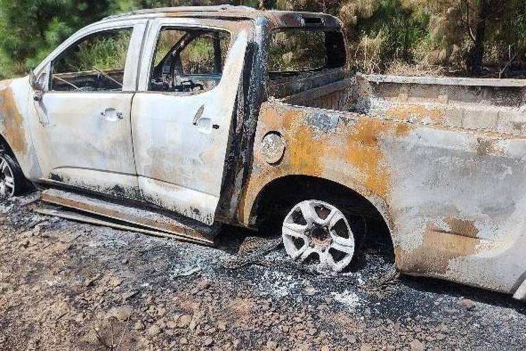 Uma caminhonete parcialmente queimada está estacionada em um terreno acidentado. A carroceria é de cor branca， apresenta manchas de ferrugem e áreas queimadas. As rodas estão danificadas.