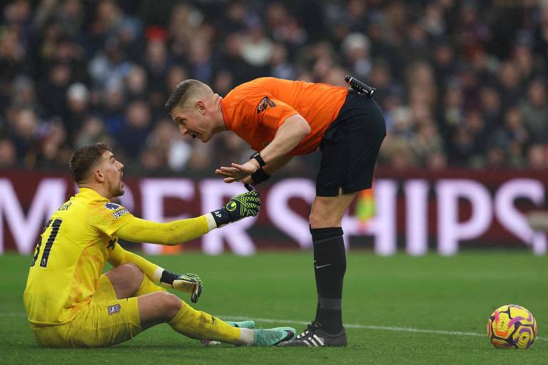 A imagem mostra um goleiro sentado no gramado， vestindo um uniforme amarelo， com uma expressão de frustração. Ele está gesticulando com a mão esquerda em direção a um árbitro， que está em pé e vestindo um uniforme laranja. O árbitro parece estar explicando algo， com a mão levantada. O fundo da imagem é desfocado， mas é possível ver uma multidão assistindo ao jogo.