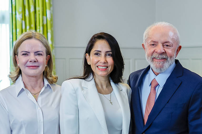 A imagem mostra três pessoas posando juntas em um ambiente interno. À esquerda， Gleisi， uma mulher com cabelo loiro e liso， vestindo uma camisa branca. No centro， Luisa Gonzalez， uma mulher com cabelo escuro e liso， usando um blazer branco e sorrindo. À direita， Lula， um homem idoso com cabelo grisalho e barba， vestido com um terno azul e uma gravata vermelha. Ao fundo， há cortinas verdes com padrões.