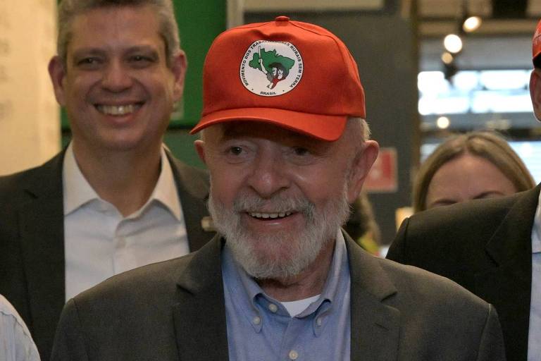 Um homem idoso， com barba e cabelo grisalho， está sorrindo e fazendo sinal de positivo com as duas mãos. Ele usa um paletó escuro e uma camisa azul clara， além de um boné vermelho com um emblema. Ao fundo， há outras pessoas sorrindo.