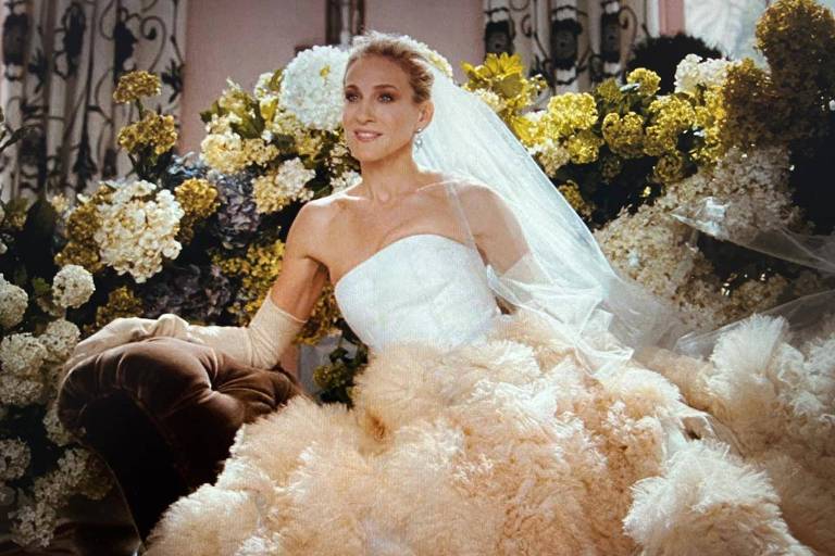 A imagem mostra uma mulher sentada em um sofá, vestindo um vestido de noiva volumoso e elegante, com uma parte superior branca e uma saia cheia de detalhes em tons claros. Ela usa um véu e tem flores no cabelo. Ao fundo, há um arranjo de flores em várias cores, criando um ambiente romântico.