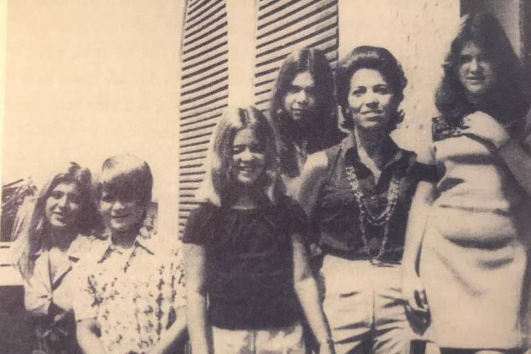 Foto em preto e branco mostra uma mulher adulta ao lado de cinco adolescentes， um deles um menino. Eles estão na área externa de uma casa e usam roupas sociais