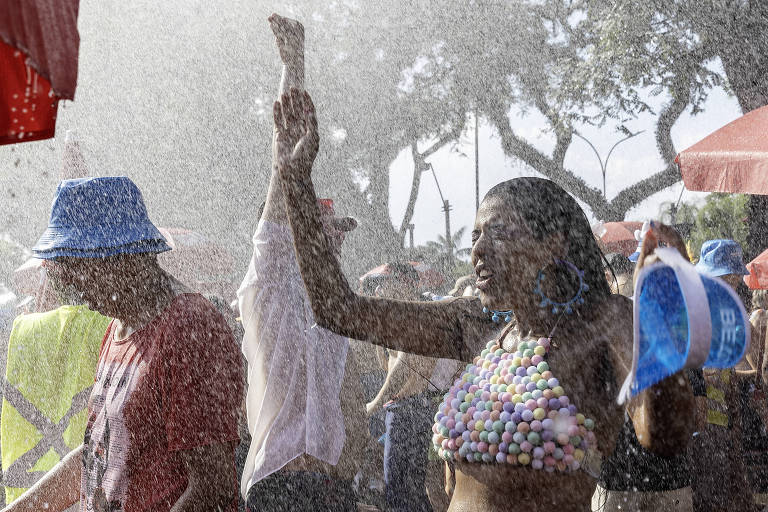 A imagem mostra um grupo de pessoas se divertindo em uma festa ao ar livre， com água sendo borrifada sobre elas. Uma mulher à frente， usando um top colorido feito de bolinhas， levanta uma das mãos enquanto sorri. Ao fundo， outras pessoas também estão se divertindo， algumas usando chapéus. O ambiente é ensolarado e festivo， com árvores e tendas visíveis.