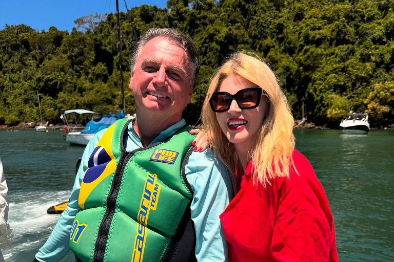 Um homem e uma mulher estão em um barco. O homem usa um colete salva-vidas verde com detalhes em amarelo e azul, e uma camisa de manga longa azul clara. A mulher está vestindo uma roupa vermelha e óculos escuros. Eles estão sorrindo e posando juntos, com um fundo de água e vegetação ao redor.