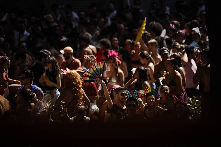 A imagem mostra uma grande multidão reunida em um evento festivo ao ar livre. As pessoas estão vestidas de forma colorida e alegre, algumas usando acessórios como leques e orelhas de coelho. A cena é iluminada por luz natural, com sombras projetadas, criando um contraste entre as áreas iluminadas e as mais escuras. A atmosfera é vibrante e cheia de energia.