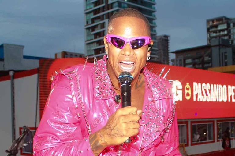 Um artista se apresenta em um palco, vestindo um traje rosa brilhante e óculos escuros. Ele segura um microfone e parece estar animado, cantando para uma multidão. Ao fundo, há prédios e uma grande quantidade de pessoas assistindo ao show.