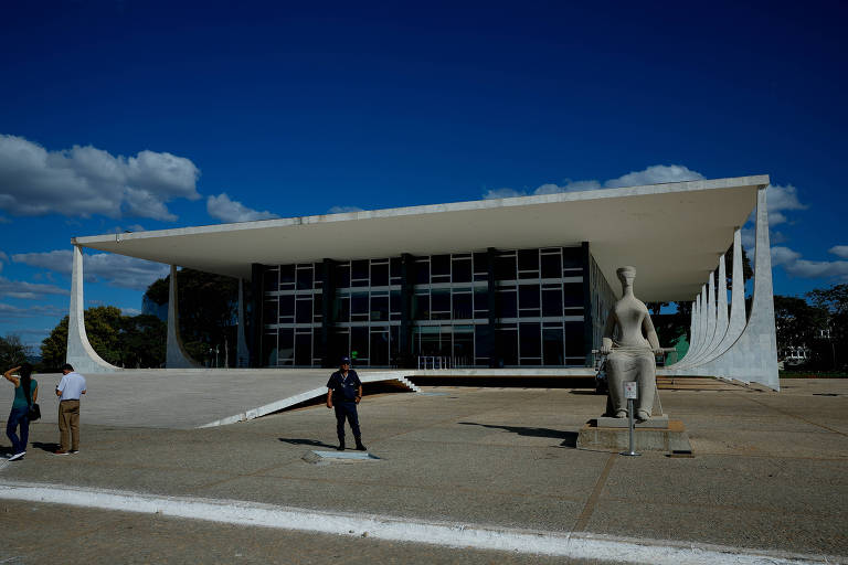 Imagem do Supremo Tribunal Federal， um edifício moderno com uma estrutura de concreto e vidro. Na frente， há uma escultura de uma figura feminina e algumas pessoas caminhando. O céu está claro com algumas nuvens.