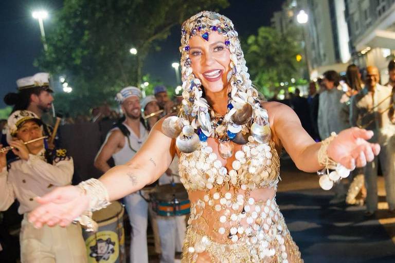 A imagem mostra uma mulher dançando em um desfile de carnaval, vestindo uma fantasia brilhante feita de conchas e lantejoulas. Ela está sorrindo e com os braços abertos, cercada por músicos que tocam instrumentos como saxofone e percussão. O ambiente é festivo, com luzes e pessoas ao fundo, criando uma atmosfera animada.