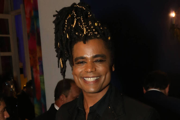 Um homem sorridente está posando em um evento social. Ele usa um traje preto com detalhes e calças listradas. O fundo é um ambiente iluminado, com pessoas e mesas ao redor.