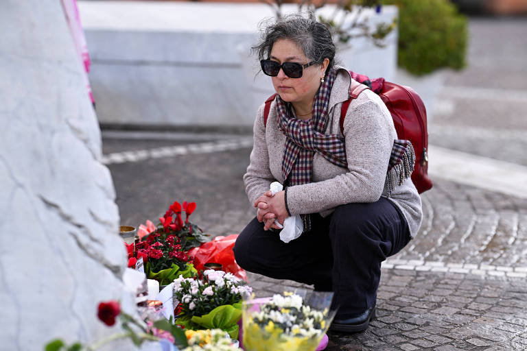 Uma mulher de cabelos grisalhos， usando óculos escuros e uma mochila vermelha， está agachada em frente a um memorial. Ela observa um local onde há flores e velas， demonstrando um momento de reflexão ou luto.