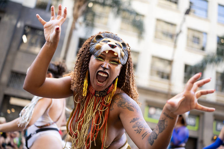 Uma mulher com cabelo cacheado e adornos coloridos, incluindo uma máscara de leão, está fazendo uma expressão intensa e dançando em um ambiente urbano. Ela usa colares e tem tatuagens visíveis nos braços. Ao fundo, outras pessoas também estão dançando, e há edifícios e árvores ao redor.